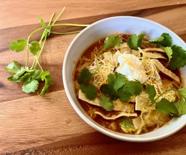 A steaming bowl of chicken tortilla soup garnished with avocado, sour cream, cheese, homemade tortilla strips, and cilantro, showcasing vibrant colors and textures, perfect for a cozy meal