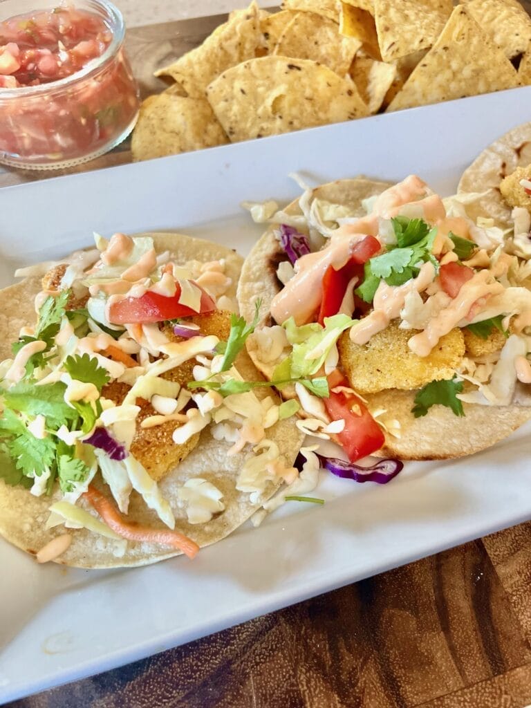 A plate of crispy, golden-brown fish tacos topped with vibrant cabbage slaw, creamy fish taco sauce, and garnished with fresh lime wedges. The tacos are served alongside tortilla chips, guacamole, and a refreshing drink, showcasing a delicious and inviting meal perfect for family dinners or gatherings.